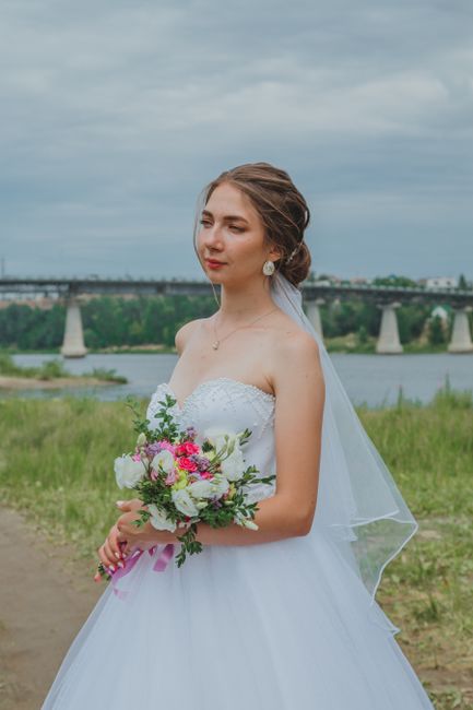 A bride dressed in a strapless white wedding gown holds a vibrant bouquet of fresh flowers, including pink roses, white peonies, and greenery, wrapped with pink ribbon. She stands outdoors on a grassy