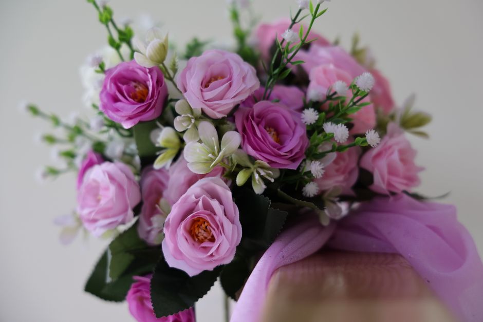 A close-up of a floral bouquet featuring soft pink and purple roses, small white daisies, and green foliage, arranged in a compact, rounded style. The bouquet is wrapped with a sheer pink ribbon, and 