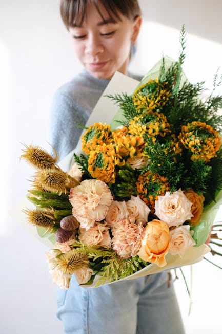 A young person with short brown hair and a gentle expression holds a large, vibrant floral bouquet in front of a neutral background. The bouquet features a mix of fresh flowers including soft pink car