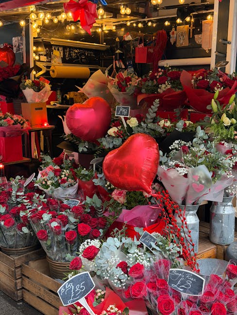 A vibrant display of floral arrangements at a flower shop, featuring numerous bouquets predominantly in shades of red, pink, and white. The scene includes heart-shaped helium balloons, some in glossy 