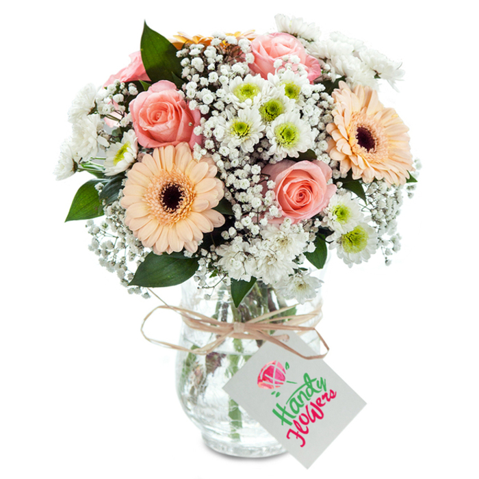 Elegantly positioned on a sunlit white surface, this handcrafted bouquet from a Lea Bridge florist captures both local character and classic beauty. Softly blushing peach-pink roses, their petals delicately unfurling, create a gentle romantic core, infusing the entire composition with sophistication. Flanking these are striking gerbera daisies in shades of ripe peach, their bold forms and chocolate-brown centers radiating warmth reminiscent of the welcoming ambience found in Lea Bridge's shops and cafés. Interspersed between these hero blooms are clouds of pure white gypsophila, each tiny floret adding a gossamer, almost ethereal quality reminiscent of English spring mornings by the River Lea. The bouquet's lush texture is deepened by clusters of white chrysanthemum blossoms, each accented by a green button center, giving an impression of dewy freshness and lively contrast. Sparse but purposeful eucalyptus leaves add structure and an invigorating, leafy note, echoing the natural beauty of Hackney Marshes nearby. Stems are artfully gathered in a transparent, lightly ribbed glass vase, revealing both the vitality of each flower and the clarity of fresh water. Around the vase's neck, a hand-tied twine ribbon bears a simple white tag-blank and inviting for personalized wishes. The thoughtful interplay of colour, form, and texture speaks to the expertise of local Lea Bridge florists and their understanding of community tastes. Perfect for gifting, celebrations, or brightening a room in E10 or along the Lea Valley, this bouquet delivers a sense of quiet celebration rooted in heartfelt local tradition.