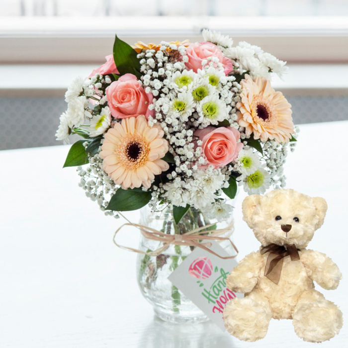 A tender anniversary-themed composition is displayed on a pristine white surface, glowing under gentle, diffused natural light that could easily be drifting in from a flat near Lea Bridge station. At the centre is a rounded bouquet settled in a clear glass jar-style vase, overflowing with pastel flowers. Pale pink roses with softly curling petals lend romantic sweetness, while peach-hued gerbera daisies, each with a distinct dark brown centre, add a playful, celebratory touch. Between them are white chrysanthemums, whose petals surround pale lime-green centres, and generous sprigs of airy white baby's breath that seem to float among the larger blossoms, softening every edge. The stems are visible inside the transparent vase, neatly gathered, and at the top a length of natural-coloured twine is tied in a simple bow. From this rustic detail hangs a slender white rectangular tag, decorated with a pink heart-like motif and elegant green lettering that hints at a handmade, thoughtfully curated gift rather than something mass-produced. To the right of the vase, slightly turned toward the viewer, a plush teddy bear sits upright. Its fur is a warm cream, textured and fluffy, inviting the thought of touch. The bear's small, dark brown button eyes, matching nose, and subtle stitched mouth give it a gentle expression, and a dark brown satin ribbon tied neatly around its neck completes its classic look. The background is softly blurred and light, with no sharp details, so that all attention rests on the flowers and bear. Together, they create an image that speaks of care, affection, and the wish to make someone in Lea Bridge feel cherished on an anniversary or special day.
