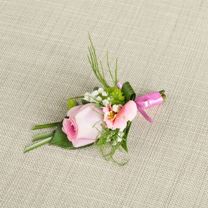 Expertly arranged on a natural, off-white woven fabric that recalls the artisanal markets of Lea Bridge, this boutonniere is an ode to thoughtful local floristry. The design centres on a blush pink rosebud, petals wound in a gentle spiral and lightly dusted with a gradient from deep rose to peachy cream, showcasing its freshness and quiet elegance. A partner blossom in blush and peach, likely an alstroemeria, offers subtle yellow notes and delicate lines-its markings reminiscent of the delicate reeds along nearby waters. Wisps of baby's breath, airy and white, interlace with vibrant green cushion chrysanthemums, providing both contrast and intricate detail. Supportive dark green leaves, elegantly folded and layered, form the base, while fine fern-like sprigs arch over the top, contributing local greenery reminiscent of the Lea Valley. The whole ensemble is bound at the stems with a lustrous, fuchsia ribbon, fastened into a crisp bow, lending an air of festivity suitable for weddings, proms, and special Lea Bridge gatherings. Gentle sunlight brings out the harmonious blend of colours and textures, making this boutonniere an ideal choice to celebrate love and community.