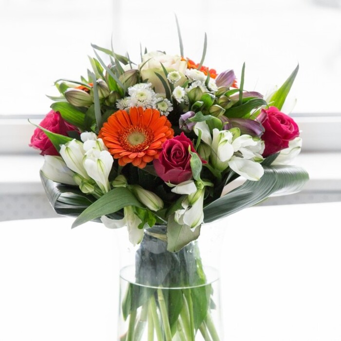 This lavish bouquet is a celebration of Lea Bridge's contemporary style and natural abundance. Central to the display are four gerbera daisies-two shining in vivid orange and two in serene, creamy peach-each radiating charm from their deep brown centres. They are gracefully complemented by magenta roses, elegantly spiralled and lush, their velvety petals offering a rich sense of romance and tradition. White alstroemeria blooms and clusters of delicate multi-petaled spray roses interlace the bolder flowers, bringing lightness and a subtle, uplifting scent. Gentle touches of blush pink and airy white filler blooms enhance the arrangement's fullness, evoking the feel of a spring afternoon by the riverbank. The entire composition is hugged by glossy, dark green leaves, with artistically curved aspidistra forming a supportive frame reminiscent of the leaf-strewn paths around Millfields Park. All of this is presented in a transparent, slender glass vase, through which the tangled green stems add to the organic elegance. Designed for celebrations, new beginnings, or as a thoughtful gesture, this bouquet resonates with the warm, inclusive atmosphere of Lea Bridge, showcasing the expertise of florists who understand the area's unique rhythm and beauty.