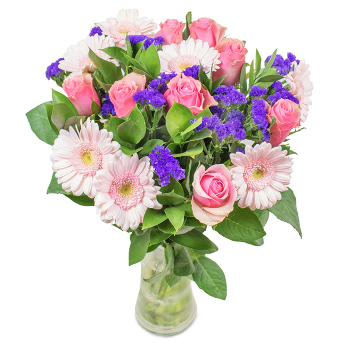 A vibrant, artfully arranged floral bouquet stands proudly in a clear glass vase, set against a luminous white backdrop-an image evocative of the airy spaces and garden views typical of Lea Bridge residences. The centerpiece features approximately 7-9 classic pink roses, their petals tightly coiled or beginning to reveal the iconic spiral, each bloom a testament to refined romance. These are beautifully complemented by 5-7 blush gerbera daisies, known for their large, sunlit faces and nearly white centres reminiscent of the fresh dawn breaking over Lea Bridge Marshes. Interspersed are vivid purple statice clusters that add a striking contrast and unique texture, mirroring the wildflower diversity along the banks of the River Lea. The deep green foliage provides a lush, abundant base, enhancing both vibrancy and freshness, as sturdy stems extend visibly through the vase's clear water. This arrangement, with its harmonious blend of pink roses, cheerful gerbera daisies, and purple accent flowers, is ideal for celebrating birthdays, anniversaries, or simply bringing stylish joy into offices and homes across the Lea Bridge community. Each element has been selected for beauty and sentiment, resulting in an arrangement that communicates celebration, affection, and artistry rooted in the local landscape.