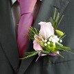 There's a subtle, heartfelt radiance in this Lea Bridge buttonhole, its beauty captured through a thoughtful arrangement of textures and hues. The soft pink rose sits prominent, its petals gently spreading, embodying the warmth and optimism felt at local celebrations from E5 to the edge of Walthamstow Marshes. A smaller pink bloom, resembling a dainty alstroemeria with purple-streaked petals, is nestled close by-each flower chosen for its expressive quality. The boutonniere is enriched by sprays of baby's breath, their delicate white fluff lending a sense of quaint romance, and vivid green from a button chrysanthemum nods to the verdant parks and waterways nearby. Wispy greens and slender leaves fan around the blooms, creating a natural grace against the dark lapel of a formal suit and a striking magenta pink tie. The look is brought together by a shimmering lavender ribbon, tied in a petite bow, reinforcing the spirit of celebration. Crafted for moments of meaning-be it a riverside wedding or a milestone event on Lea Bridge Road-this floral accent reflects the careful work and community pride of Lea Bridge's local florists, making special occasions even more memorable.